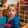A young toddler, approximately 2-3 years old, sits at a small table in what appears to be a daycare or preschool classroom. The child is wearing a blue hoodie with rainbow patterns over a yellow shirt and is looking directly at the camera. The background shows well-organized shelving units filled with art supplies, toys, and educational materials.