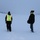 Two individuals are standing in a snow-covered open area during an active snowstorm with low visibility conditions. One person is wearing a high-visibility safety vest over winter clothing, while the other wears a dark jacket with a yellow collar and a black beanie. Both appear to be observing or surveying the surrounding landscape, with a treeline and what appears to be a structure or vehicle faintly visible in the background.