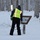 A person wearing a high-visibility safety vest and winter clothing is leaning over an informational trail sign in a snow-covered forest. The sign appears to read 'Chena River Nature Trail,' suggesting this is likely in Alaska. The scene depicts a winter environment with heavy snow accumulation on the ground and trees.