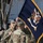 Kentucky Guardsmen Assume Authority A military ceremony is taking place outdoors, featuring U.S. Army soldiers in OCP (Operational Camouflage Pattern) uniforms handling a dark navy blue Infantry unit guidon or colors with gold fringe. The flag displays the U.S. Army Infantry insignia with an eagle and the word 'INFANTRY' visible. Multiple soldiers are assembled in formation in the background, suggesting a formal change of command or colors ceremony.