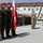Kentucky Guardsmen Assume Authority A military ceremony or parade formation featuring Polish and American soldiers standing at attention on a gravel surface in front of modular military buildings. Polish soldiers in distinctive camouflage uniforms and red berets are prominently positioned on the left, holding the Polish national flag (white and red with the Polish eagle emblem). American soldiers in MultiCam/OCP uniforms are visible on the right, holding what appears to be a unit guidon flag.