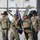 Kentucky Guardsmen Assume Authority A U.S. Army change of command or guidon ceremony is taking place outdoors on a gravel surface, likely at a forward operating base. Two soldiers in the center are exchanging a blue unit guidon/flag, while two other soldiers wearing distinctive black cavalry Stetson hats stand at attention in the background. All personnel are wearing OCP (Operational Camouflage Pattern) uniforms with U.S. Army insignia.