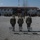 Kentucky Guardsmen Assume Authority A military ceremony is taking place at what appears to be a multinational military base or forward operating base. Three soldiers stand with their backs to the camera facing a formation of troops, with an American flag and at least one other flag visible at the center. Multiple national flags are displayed on the building in the background, suggesting a NATO or coalition operation.