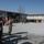 Kentucky Guardsmen Assume Authority A multinational military ceremony is taking place in an outdoor courtyard of what appears to be a forward operating base or military installation. Soldiers in camouflage uniforms are standing in formation and saluting, with multiple national and organizational flags visible including Polish, NATO, EU, and Turkish flags. The setting includes modular/prefabricated buildings typical of temporary military installations.