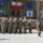 Kentucky Guardsmen Assume Authority A military formation ceremony is taking place at what appears to be a Polish military installation, with soldiers in US Army MultiCam/OCP uniforms standing in formation. Three large flags are displayed on the building behind them: NATO, Poland, and the European Union. Unit banners and signs in Polish are also visible, suggesting a joint or allied military event.