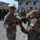 Kentucky Guardsmen Assume Authority A military ceremony is taking place outdoors at what appears to be a forward operating base or military installation. Two soldiers in the foreground are handling a folded military guidon or colors with gold fringe, while a formation of soldiers stands at attention in the background. Patches visible on one soldier indicate KFOR (Kosovo Force) and RC-East affiliation.