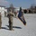 Kentucky Guardsmen Assume Authority A military ceremony is taking place on a gravel parade ground at what appears to be a forward operating base or military installation. Two soldiers in the foreground are holding a dark blue unit guidon/flag with gold fringe and insignia, while additional personnel are visible in the background. The setting includes modular/containerized buildings typical of a deployed military environment.