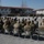 Kentucky Guardsmen Assume Authority A military ceremony is taking place at an outdoor parade ground of what appears to be a multinational military base. Seated soldiers in various camouflage uniforms and colored berets face formations of standing troops in the background. Flags including the Polish flag and NATO flag are visible on the building facade, indicating a multinational military installation.