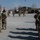 Kentucky Guardsmen Assume Authority A military formation ceremony is taking place on a gravel parade ground at what appears to be a forward operating base or military installation. Soldiers in OCP (Operational Camouflage Pattern) uniforms are standing in formation on both sides, observing a central ceremony involving color guards and flag bearers. The setting includes modular/temporary military buildings and a flagpole in the background, consistent with a deployed or overseas base environment.
