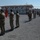 Kentucky Guardsmen Assume Authority A military ceremony or parade is taking place on a gravel parade ground at what appears to be a military base or camp. Soldiers in camouflage uniforms and green berets are standing in formation holding various unit flags and guidons. The setting includes modular/prefabricated white buildings with red roofs in the background.