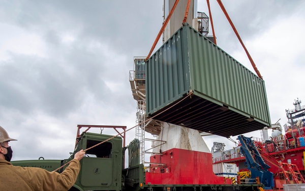 A military loading operation is underway at a port facility, where a green shipping container is being lifted by crane from or onto a military olive-drab heavy truck. A Navy sailor wearing a tan jacket, hard hat, face mask, and camouflage pants is guiding the operation using a tag line rope. A large red vessel is docked in the background, consistent with a research or support ship.