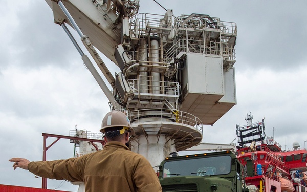A military or naval personnel member wearing digital camouflage utilities and a hard hat is directing a military tactical truck (appears to be an LVSR or similar heavy military vehicle) at a port facility. The scene takes place on a dock alongside a large red-hulled vessel with a massive crane structure visible overhead. The setting suggests a military logistics or equipment loading/offloading operation.