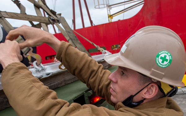 A worker wearing a tan/beige hard hat with a green 'Safety Is My Responsibility' sticker is performing a hands-on task involving straps or tie-down hardware on industrial equipment. He is wearing a brown fleece jacket and has a communication headset around his neck. The setting appears to be an outdoor industrial or maritime environment, with a red structure visible in the background.