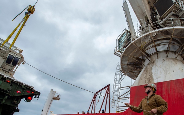 A military service member in Navy working uniform and hard hat guides a large military vehicle being lifted by crane onto or off of a cargo ship. The red-hulled vessel visible in the background is named 'PICASSO NASSAU' and appears to be a roll-on/roll-off (RoRo) or heavy-lift cargo ship docked at a port. The operation appears to be a military equipment loading or offloading exercise at a pier.