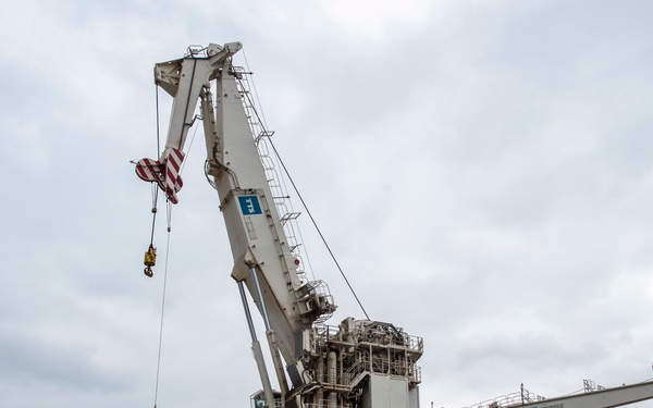 A large industrial port crane is in operation at a commercial or naval dock facility. A red vessel named 'PICASSO NASSAU' is moored alongside the quay, with cargo and crates visible on the dock. Several personnel are standing on the dock observing or supervising the crane's lifting operation under an overcast sky.