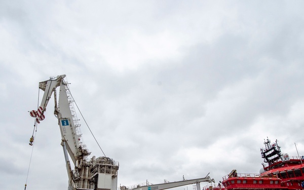 A large red offshore support vessel named 'UltraDeep' is docked at a port facility, alongside a massive white pedestal crane labeled 'Picasso Nassau'. The scene shows active port operations with personnel on the dock and cargo/equipment staged nearby. The overcast sky and wide-angle perspective emphasize the industrial scale of the vessels and equipment.