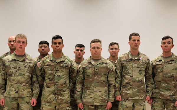 A group of approximately ten U.S. Army soldiers in OCP (Operational Camouflage Pattern) uniforms posing for a formal group photograph indoors. They are standing in a single row against a white wall in what appears to be a conference or briefing room. Name tapes and rank insignia are visible on several uniforms.