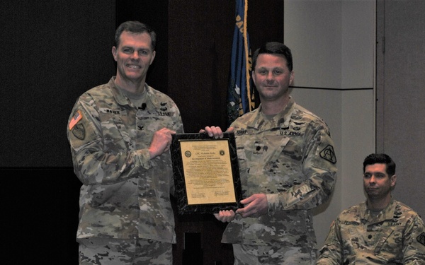 Two U.S. Army soldiers in OCP (Operational Camouflage Pattern) uniforms are posing together while holding a framed plaque or certificate during what appears to be an awards or recognition ceremony. A third soldier is seated in the background observing the presentation. The setting appears to be an auditorium or conference room with a military flag visible in the background.