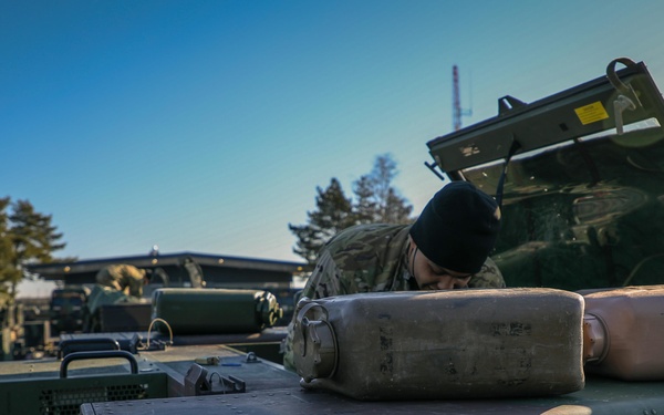 A military service member wearing camouflage uniform and black beanie is inspecting or working inside an open compartment of a military vehicle. The foreground shows the top surface of a green armored military vehicle with stenciled markings including 'NO STEP' and two metal containers resting on top. Multiple additional military vehicles are visible in the background at what appears to be a military staging or motor pool area.