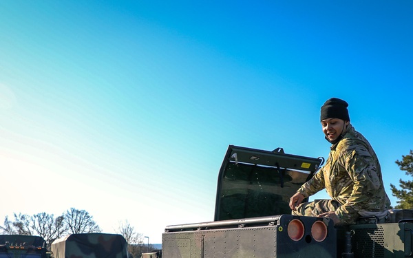 A soldier in camouflage uniform and black beanie hat is working on top of a military vehicle, appearing to inspect or operate equipment. The scene takes place in an outdoor military staging area with multiple military vehicles visible in the background. The setting appears to be a European military installation based on the vehicle markings and surrounding environment.