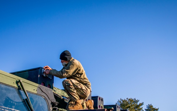 A military personnel member in olive/tan combat uniform and black beanie is working on top of a military vehicle, appearing to perform maintenance or equipment checks. The scene shows a motor pool or staging area with multiple military vehicles lined up under a clear blue sky. Camouflage netting is visible in the foreground, and the partial text 'MOB' is visible on the vehicle's side.