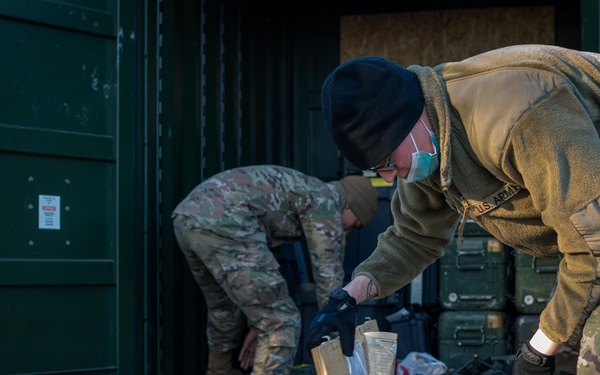 Two U.S. Army soldiers in MultiCam/OCP uniforms are handling wooden military shipping crates outdoors near green metal storage containers. The foreground soldier, wearing a black beanie and face mask, is inspecting or opening a labeled wooden crate marked 'A23'. The scene appears to be a logistics or equipment staging operation.