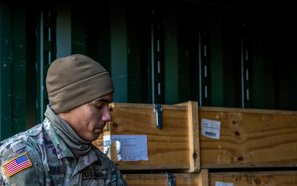 A U.S. Army soldier wearing MultiCam (OCP) camouflage uniform and a tan fleece cap is working inside or at the entrance of a military shipping container. The soldier is handling what appears to be tactical gear or a military bag/carrier. Wooden military crates with shipping labels are stacked in the background inside the container.