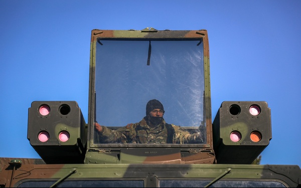 A military personnel is seated inside the enclosed turret or operator station of a military vehicle, viewed from below against a clear blue sky. The vehicle appears to be a modern military platform equipped with what appear to be infrared countermeasure or sensor pods on both sides of the enclosure. The soldier is wearing camouflage uniform and a dark balaclava or cold-weather head covering.