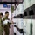 Two U.S. military personnel in OCP (Operational Camouflage Pattern) uniforms are conducting an inventory or inspection of stacked storage containers in what appears to be a military warehouse or logistics facility. One airman/soldier is scanning barcodes on the containers while the other reviews paperwork. The facility contains rows of large white and black plastic storage containers with labels and shipping documentation attached.