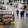 A large military logistics warehouse showing personnel in camouflage uniforms and civilians working with palletized cargo. Multiple pallets are loaded with boxes labeled 'SURE-Pak' and secured with cargo netting and straps. The scene depicts an active military supply or humanitarian aid operation.