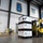 A warehouse facility identified as 'The Superport' shows military logistics operations in progress. A yellow Hyster forklift operated by a civilian worker is transporting a stacked pallet of white boxes labeled 'GSA Contract Holder'. Military personnel in camouflage uniforms are visible on the warehouse floor managing strapping materials.