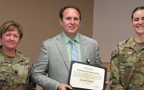 Three individuals pose for a formal photo, with a civilian man in the center holding a Certificate of Appreciation issued to 'Matthew Crumpton.' He is flanked by two U.S. Air Force personnel in OCP (Operational Camouflage Pattern) uniforms. The setting appears to be an indoor military or government facility.
