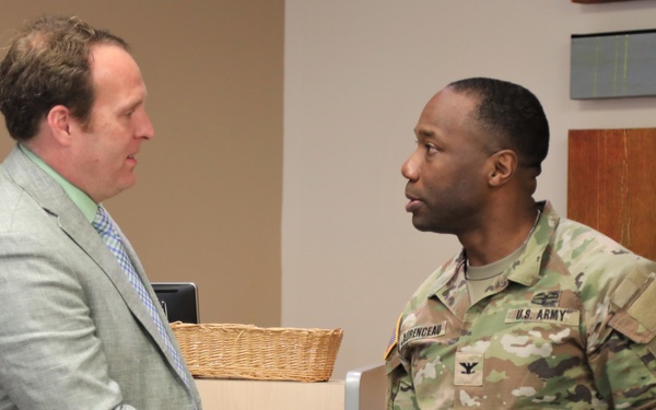 A civilian man in a gray blazer and plaid tie is shaking hands with a U.S. Army soldier in OCP (Operational Camouflage Pattern) uniform. The soldier's name tape reads 'LORENCEAU' and displays a Colonel's eagle insignia, indicating the rank of Colonel. The setting appears to be an indoor facility, possibly a military or government office.