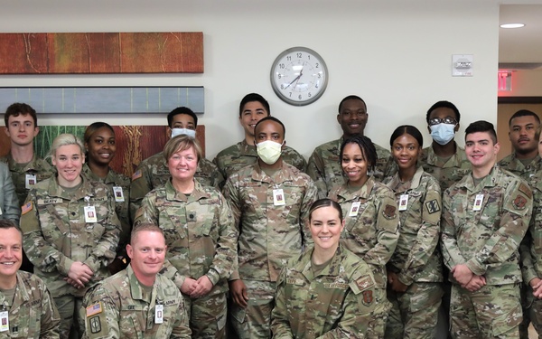 A large group of approximately 20 military personnel in U.S. Army/Air Force OCP (Operational Camouflage Pattern) uniforms pose for a formal group photo indoors. One civilian in a green suit and tie is present among the uniformed personnel. The setting appears to be a medical or administrative facility, suggested by the interior decor, ID badges, and at least two individuals wearing face masks.