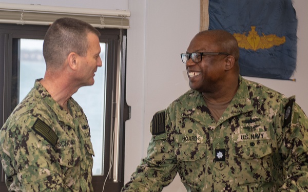 Two U.S. Navy personnel in NWU Type III (woodland digital camouflage) uniforms are shaking hands in what appears to be an office or administrative space. The individual on the right, identified by name tape as 'HOARD,' is smiling broadly and wears a Lieutenant Commander rank insignia (oak leaf). A U.S. Navy flag with a submarine warfare qualification device is visible in the background.