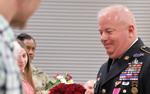 A senior U.S. Army non-commissioned officer in dress blues uniform is holding roses during what appears to be a military ceremony, likely a retirement or awards event. He is smiling and holding a bouquet of red and pink roses, with other attendees visible in the background also holding flowers. The setting appears to be inside a military facility or armory.