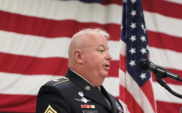 A senior U.S. Army non-commissioned officer in dress uniform is speaking at a podium during what appears to be a formal military ceremony. The individual wears the rank of Sergeant Major of the Army or Command Sergeant Major, with numerous awards and badges visible on their uniform. A large American flag is displayed prominently in the background.