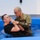A U.S. Army soldier in camouflage uniform is performing a baptism ceremony in a portable blue pool. One person is being immersed in water while the other, wearing military OCP (Operational Camouflage Pattern) uniform with a '1st Infantry Division' patch, administers the rite. This appears to be a military chaplain conducting a religious baptism ceremony.