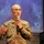 A U.S. Army soldier named Kim, wearing an OCP (Operational Camouflage Pattern) uniform, is speaking into a handheld microphone at what appears to be a religious or community event. A cross insignia on the uniform suggests the individual may be a military chaplain. The background displays a projected screen with the word 'Betania' visible.
