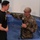 A US Army soldier in OCP (Operational Camouflage Pattern) uniform is standing in a baptismal pool holding a microphone, with one hand extended toward a young man in a black Army t-shirt. The soldier appears to be conducting or participating in a religious baptism ceremony. The setting appears to be an indoor chapel or church facility.