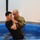 A U.S. Army chaplain in OCP (Operational Camouflage Pattern) uniform is performing a baptism ceremony in a portable blue water tank. The chaplain, who is bald and wearing glasses, is supporting a younger man dressed in a black t-shirt as he is being immersed. The setting appears to be an indoor military chapel or multipurpose facility.