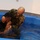 A military chaplain in U.S. Army camouflage (OCP/MultiCam) uniform is performing a baptism ceremony in a portable blue baptismal pool. He is immersing a younger individual, who is wet and bowing forward in the water. The setting appears to be an indoor military chapel or facility.