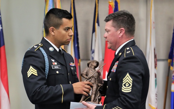 Two U.S. Army soldiers in dress uniforms are participating in a formal military ceremony. A Sergeant (name tag reads 'MORA') is receiving or presenting a bronze military figurine/statue to a Sergeant Major, as indicated by the rank insignia visible on their uniforms. The ceremony takes place in front of a display of multiple military and state flags.