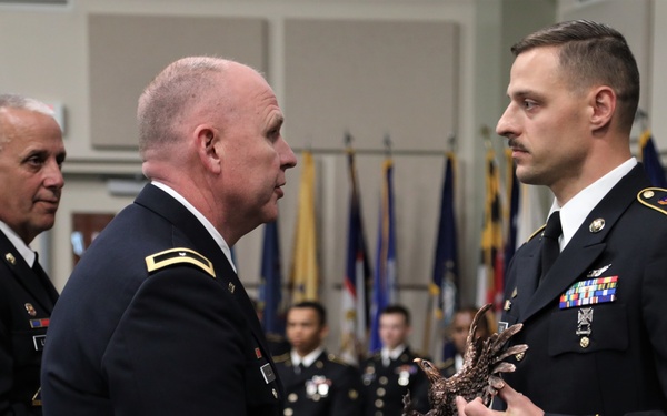 A formal U.S. Army military ceremony is taking place indoors, where a senior officer is presenting a bronze eagle trophy/award to a Staff Sergeant. Multiple service members in Army Service Uniforms (ASUs) are present, with a color guard and multiple flags visible in the background. The setting appears to be a banquet hall or conference center.