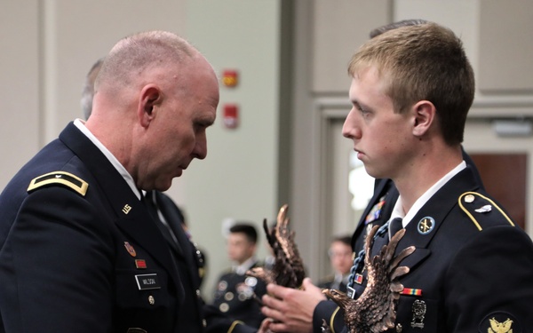 Two U.S. Army soldiers in dress blue uniforms are engaged in a formal award or trophy presentation ceremony. The senior officer on the left is handing a bronze eagle trophy to the younger soldier on the right. The event appears to take place in a banquet or conference hall with other uniformed personnel seated in the background.