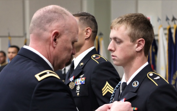 A military awards ceremony is taking place in an indoor venue. A senior officer is pinning a medal or decoration onto the uniform of a younger soldier in U.S. Army dress blues. Additional uniformed soldiers and color guard flags are visible in the background.