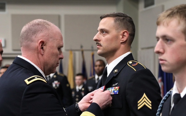 A formal U.S. Army military ceremony is taking place indoors, where a senior officer is pinning a medal or award onto a Sergeant's dress uniform. Multiple soldiers in Army Service Uniforms (ASUs) are present, standing at attention. The setting appears to be a formal event hall with military flags visible in the background.