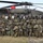 A group of approximately 18-20 military personnel pose for a group photo in front of a United States Army UH-60 Black Hawk helicopter. The group includes soldiers in US Army MultiCam (OCP) uniforms on the flanks and a larger contingent in what appears to be a different camouflage pattern, likely allied or partner nation forces, wearing full tactical gear. The photo appears to be taken outdoors on a grassy area under overcast skies.