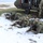Military personnel in camouflage uniforms are conducting a tactical exercise in a snowy field, having deployed from a military helicopter visible in the background. Soldiers are in prone firing positions forming a defensive perimeter, armed with various rifles and light machine guns. The scene appears to be a training exercise or simulated assault operation involving multiple personnel from what appear to be different military units.