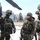 A group of heavily armed military personnel in camouflage uniforms are gathered outdoors in a winter environment near what appears to be a helicopter rotor blade visible at the top of the frame. The soldiers are equipped with full tactical gear including helmets, body armor, weapons, and communication equipment. The setting appears to be a military base or training facility with snow on the ground.